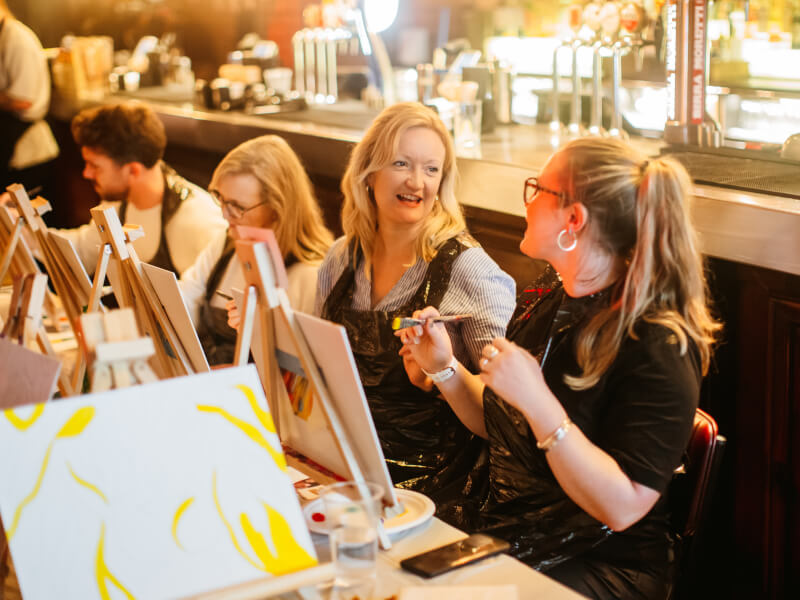 Group of people socialising and painting at a paint and sip class for team building