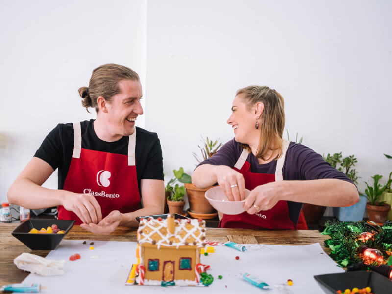 A man and woman laughing wearing aprons whilst decorating a gingerbread house.