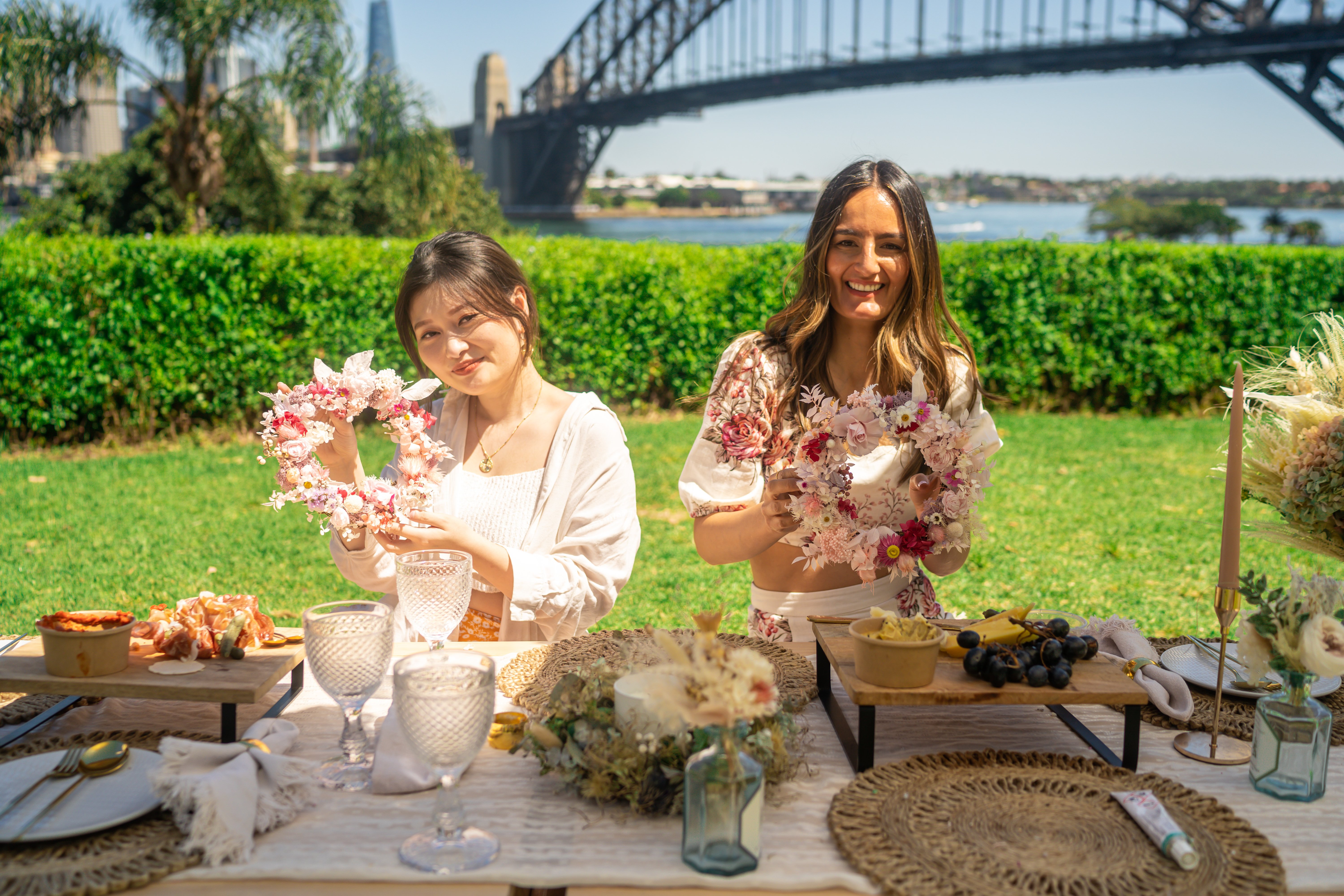 Two women holding their DIY flower crowns and sitting on a picnic outside