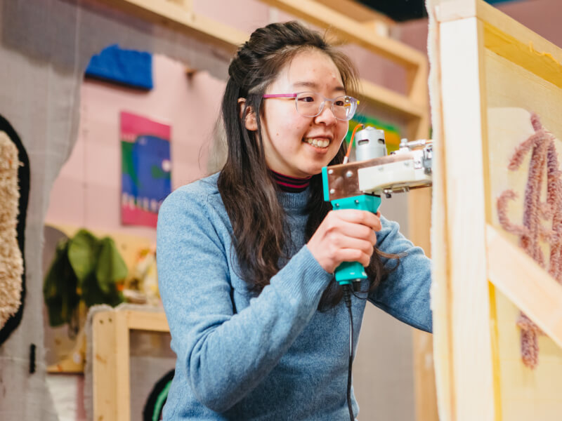 Girl holding a rug tufting gun against a frame