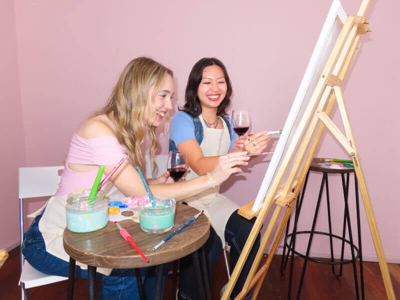 Two women painting and holding a glass of wine