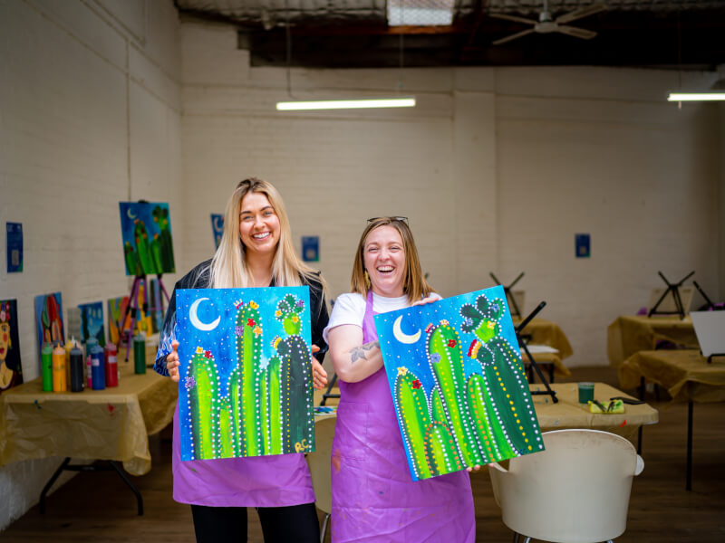 Two women smiling with their artworks at a Canberra paint and sip class