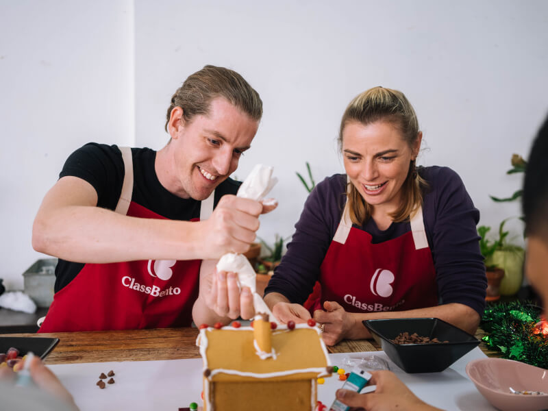 Man piping icing onto gingerbread house next to a smiling woman