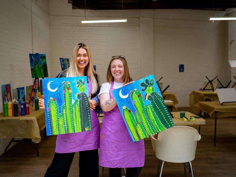 Two women posing with their finished artworks at a paint and sip party