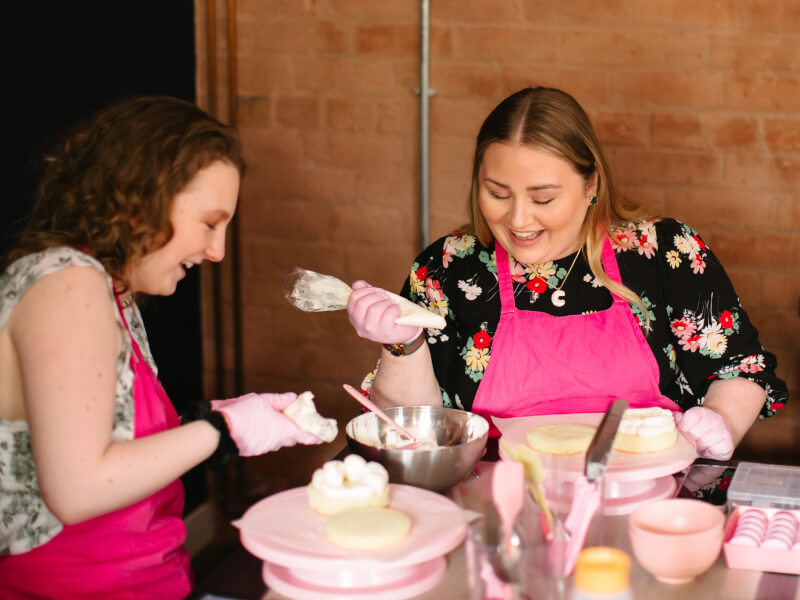 Two women laugh while they decorate cakes at a workshop