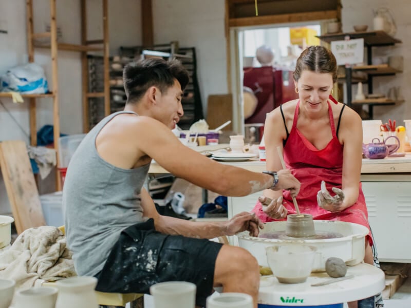 Man helping woman wearing pink apron at wheel throwing class.