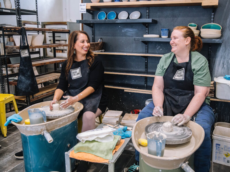 Two women smiling at a pottery wheel throwing class.