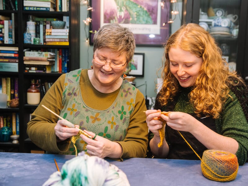 Older woman smiling and knitting with her granddaughter at a Mother's Day knitting class