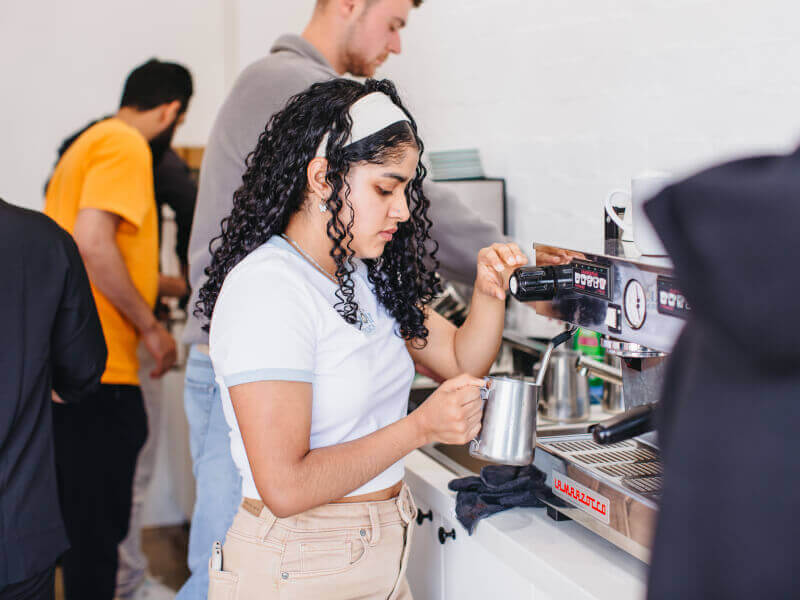 A woman practicing milk frothing at a busy barista course