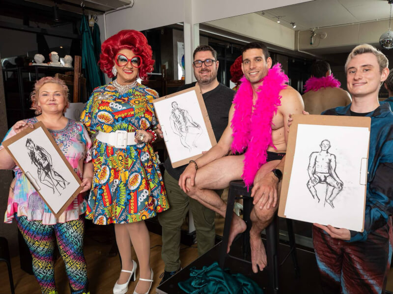 A group pose with their life drawings and male model and their drag queen teacher
