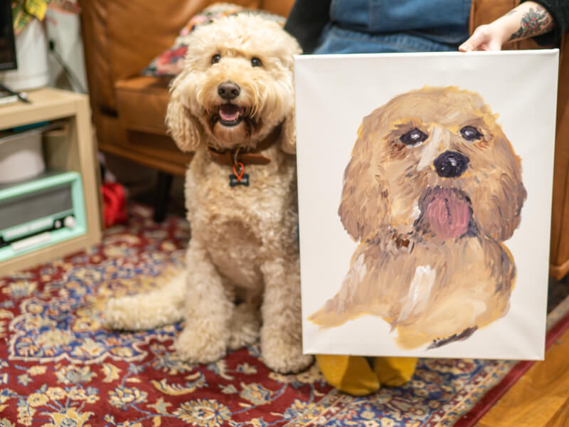 A dog sitting next to his life-like portrait on canvas