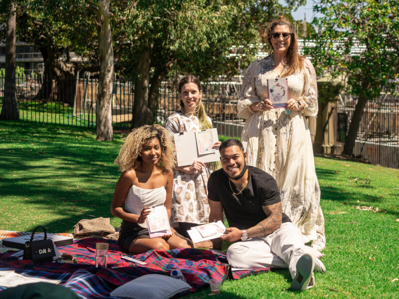 A group of friends hold their finished painting in an outdoor picnic setup