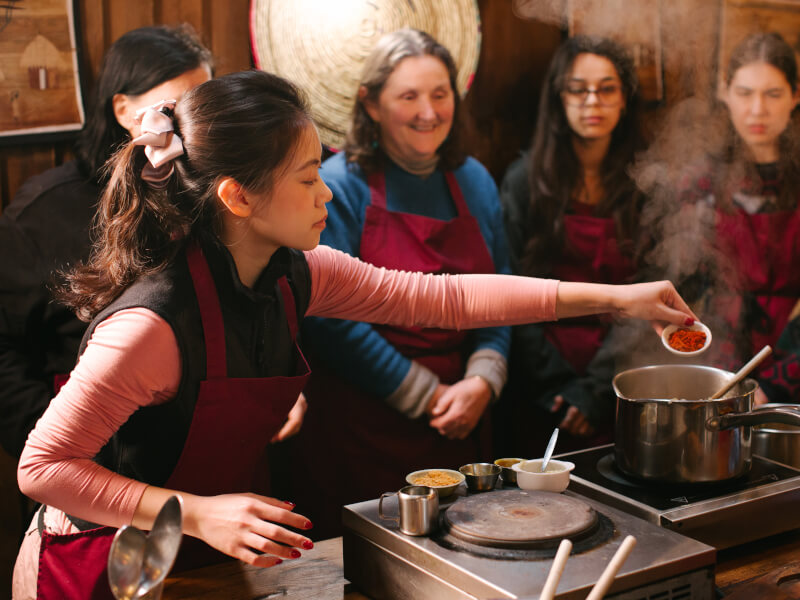 Woman adding chilli powder to pot while students watch at a Melbourne cooking class